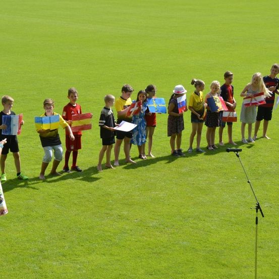 Eine Gruppe von Kindern mit verschiedenen Flaggen steht in einer Reihe auf einem grünen Feld. Einige tragen Hüte und Turnschuhe, während andere barfuß sind. Ein Mikrofon auf einem Ständer ist vorhanden.