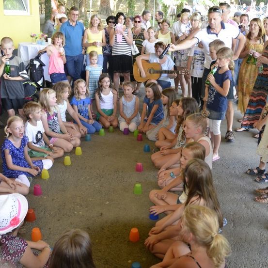 Eine Gruppe von Kindern und Erwachsenen ist in einem Kreis versammelt, einige sitzen und einige stehen, mit einem Mann, der eine Gitarre spielt und ein Mikrofon hält. Sie befinden sich im Freien, und einige haben Becher vor sich.