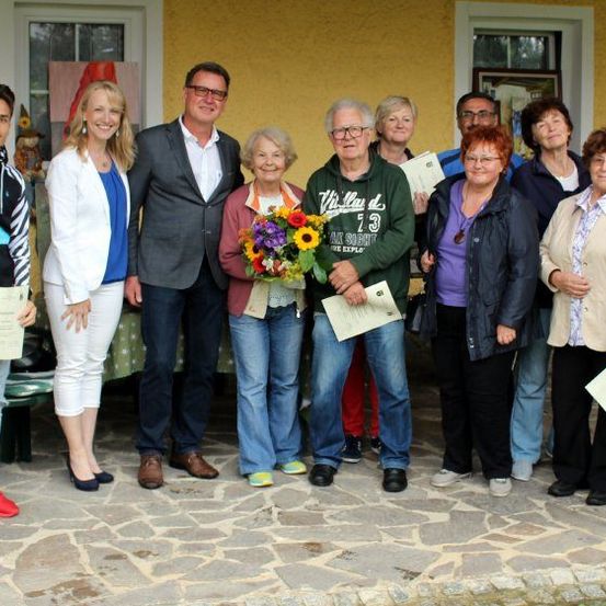 Eine Gruppe von Menschen steht draußen, möglicherweise für ein Foto, hält Urkunden und Blumen in den Händen. Hinter ihnen steht ein gelbes Haus mit Glasfenstern.