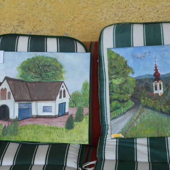 Zwei Gemälde sind auf gestreiften Kissen ausgestellt. Das linke Gemälde zeigt ein Haus mit einem braunen Dach und blauen Türen, während das rechte Gemälde eine gewundene Straße mit einer kleinen Kapelle in der Ferne darstellt.