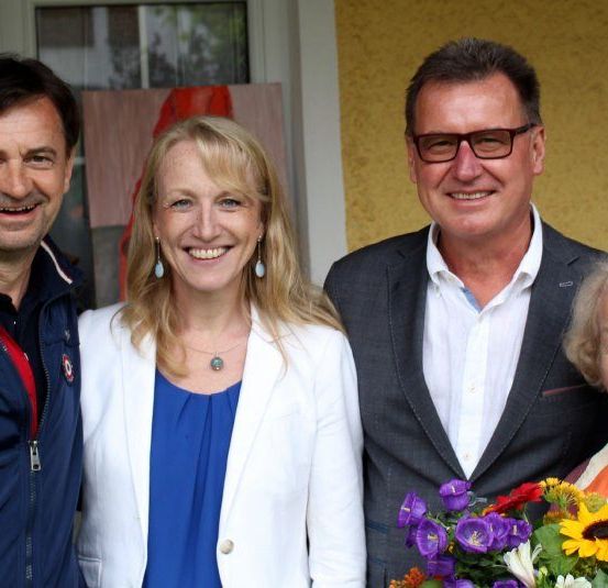 Vier Personen posieren lächelnd für ein Foto. Ein Mann in einem blauen Jackett steht links, während eine Frau in einem weißen Blazer neben ihm steht. Ein anderer Mann in einem Anzug und mit Brille steht rechts, und eine ältere Frau, die einen Blumenstrauß hält, steht neben ihm.