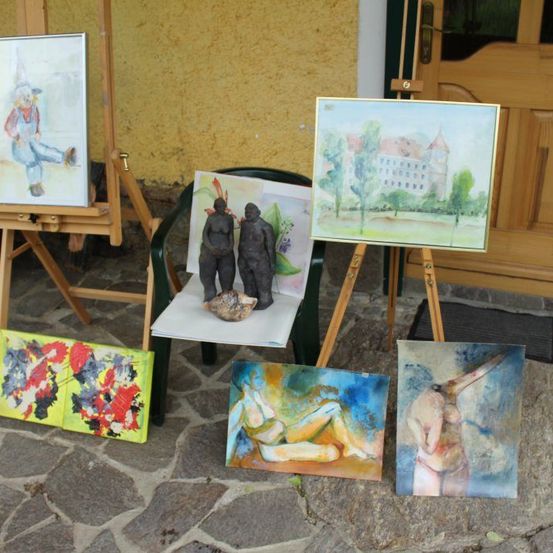 Eine Sammlung von Gemälden und Skulpturen auf Staffeleien vor einem Gebäude mit gelber Wand. Es gibt zwei Skulpturen von Menschen und ein Gemälde eines Gebäudes.
