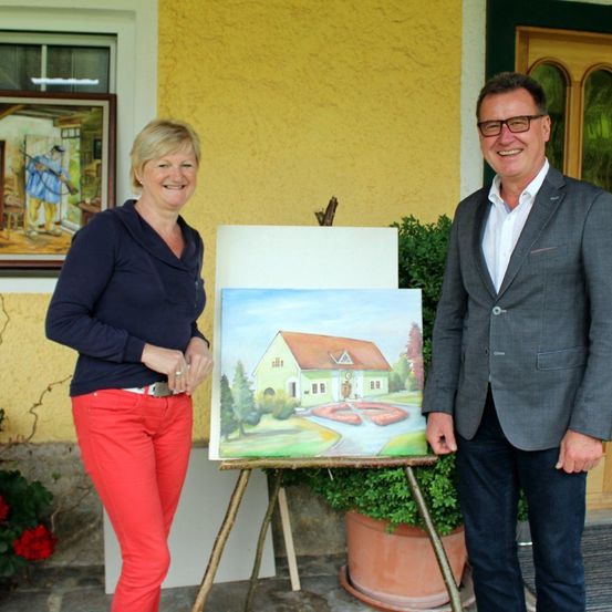 Ein Mann und eine Frau stehen neben einem Staffelei, auf dem ein Gemälde eines Hauses steht. Die Frau lächelt und trägt rote Hosen.