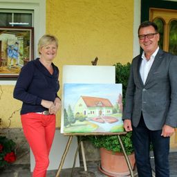 Ein Mann und eine Frau stehen neben einem Staffelei, auf dem ein Gemälde eines Hauses steht. Die Frau lächelt und trägt rote Hosen.