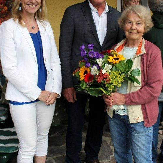 Drei Menschen stehen lächelnd im Freien. Die Frau rechts hält einen bunten Blumenstrauß. Der Mann in der Mitte trägt einen Anzug, während die Frau links einen weißen Blazer und Jeans trägt.