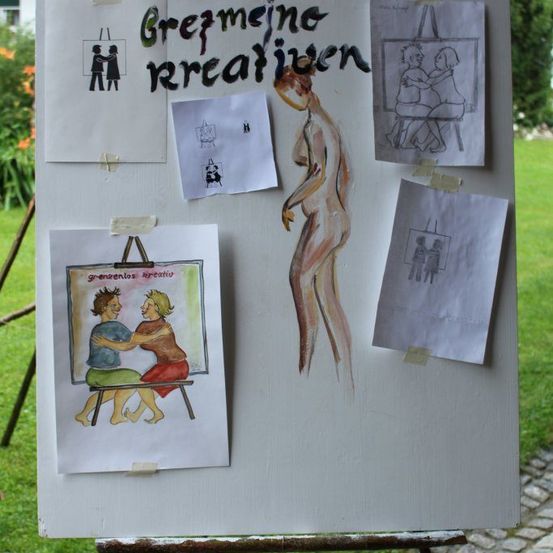 Ein weißes Brett zeigt verschiedene Zeichnungen und Skizzen, darunter ein Paar, eine nackte Figur und ein Paar, das auf einer Bank sitzt. Das Brett steht draußen, mit Gras und Pflanzen im Hintergrund.