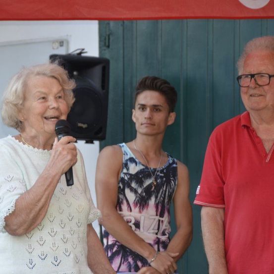 Eine ältere Frau spricht in ein Mikrofon, während sie mit einem jungen Mann und einem älteren Mann vor einer grünen Wand steht.
