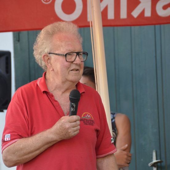 Ein älterer Mann mit Brille spricht in ein Mikrofon unter einem roten Banner. Eine Frau mit langen Haaren steht im Hintergrund, in der Nähe eines Lautsprechers.