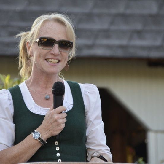 Eine Frau in traditioneller Kleidung lächelt und hält ein Mikrofon, wahrscheinlich gibt sie eine Rede bei einer Outdoor-Veranstaltung.