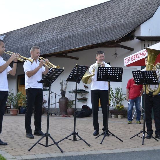 Fünf Männer stehen vor einem Gebäude und spielen Trompeten und Tuben. Sie stehen um Notenständer auf einem gepflasterten Gehweg. Dahinter stehen Topfpflanzen und ein Schild mit einem Logo. Im Hintergrund sind ein Mann in einem roten Hemd und eine weitere Person zu sehen.