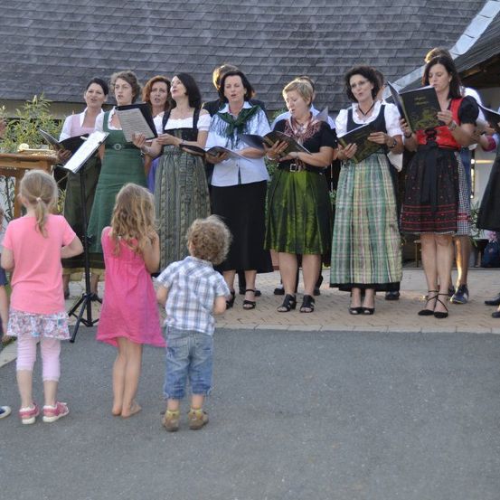 Ein Chor von Frauen in traditioneller Kleidung singt vor einer Gruppe von Kindern auf einem gepflasterten Bereich.