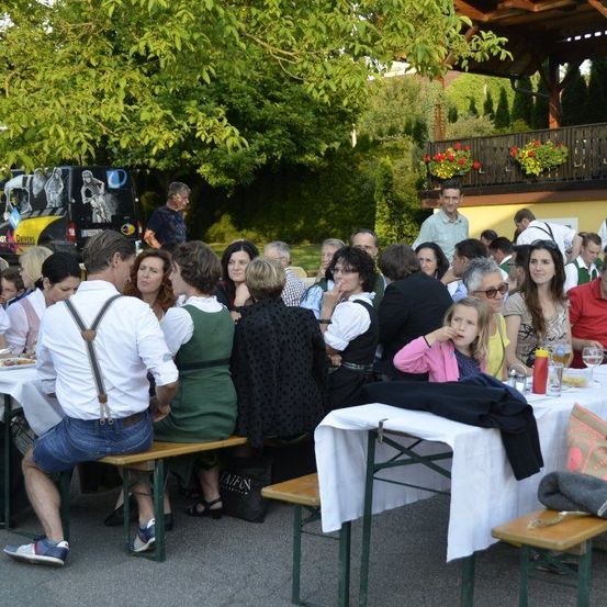 Ein Outdoor-Essevent mit Menschen an Tischen. Ein Kind isst, während andere plaudern. Im Hintergrund befinden sich Bäume und ein Gebäude mit Balkon.