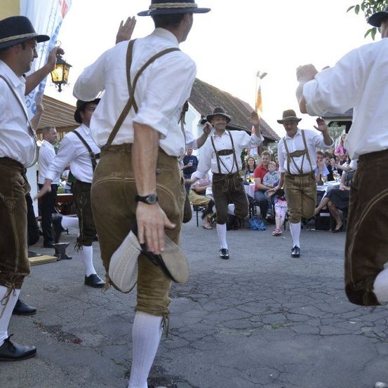 Fünf Männer in traditioneller bayerischer Kleidung führen einen Tanz vor einer Menschenmenge bei einer Veranstaltung im Freien auf.