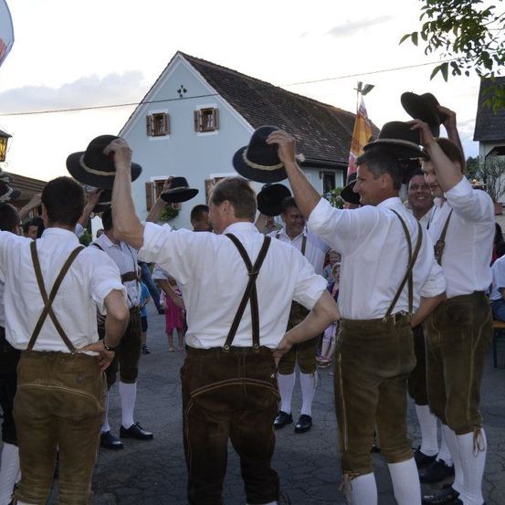 Eine Gruppe von Männern in traditioneller Kleidung, möglicherweise für ein Fest, steht im Kreis und hebt Hüte hoch. Ein Haus und eine Flagge sind im Hintergrund zu sehen.