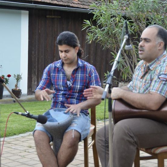 Zwei Männer sitzen im Freien auf Stühlen und treten auf. Einer spielt eine Trommel, der andere hält eine Gitarre. Beide singen in Mikrofone. Ein Gebäude und Grünpflanzen sind im Hintergrund.