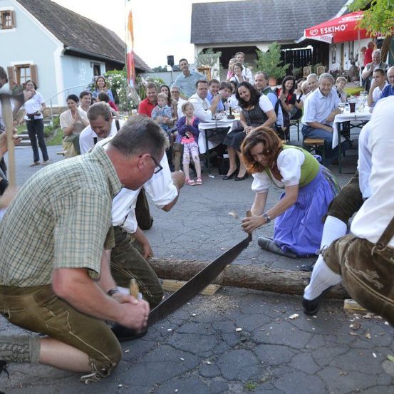 Eine Gruppe von Menschen in traditioneller Kleidung sägt einen Baumstamm auf der Straße vor einer Menge.