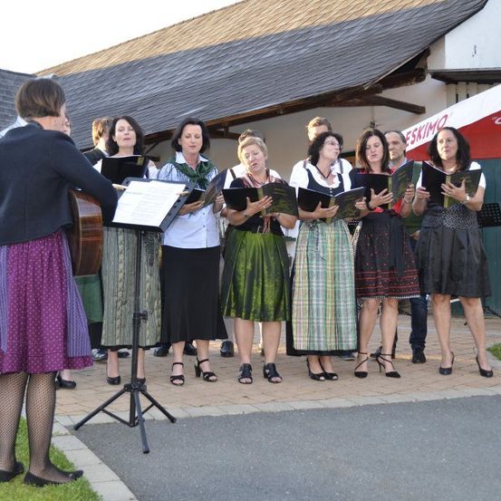 Ein Frauenchor steht auf einem Weg und singt, während sie Notenblätter halten. Eine Frau steht vor ihnen und hält eine Gitarre. Dahinter befindet sich ein Gebäude mit einem Dach.