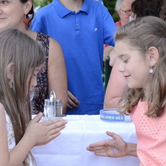Zwei junge Mädchen sitzen an einem Tisch, eines hält eine silberne Salz- und Pfeffermühle. Hinter ihnen beobachtet eine Frau in einem Blumentops, während andere im Hintergrund stehen.