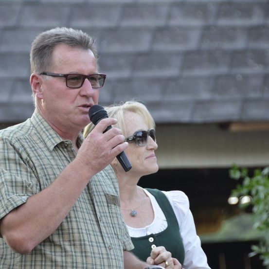 Ein Mann mit Brille hält ein Mikrofon und spricht, während eine Frau in einem grünen Kleid und Sonnenbrille hinter ihm steht. Sie sind draußen in der Nähe eines Gebäudes mit einer Steinmauer.