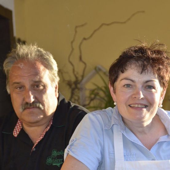 Ein Mann mit einem Schnurrbart und eine Frau mit kurzen Haaren sitzen nebeneinander und lächeln. Der Mann trägt ein schwarzes T-Shirt mit einem grünen Logo, und die Frau trägt ein blaues Hemd und eine weiße Schürze. Hinter ihnen ist eine gelbe Wand.