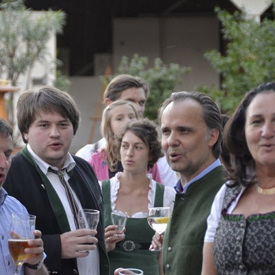 Eine Gruppe von Menschen bei einer gesellschaftlichen Veranstaltung. Ein Mann und eine Frau halten Getränke, während andere zusehen. Sie sind im Freien in der Nähe von Pflanzen und einem Gebäude.