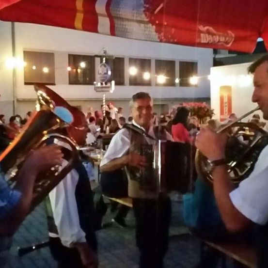 Mehrere Personen spielen Instrumente unter einem roten Regenschirm in einer Außenumgebung. Eine Menge beobachtet.