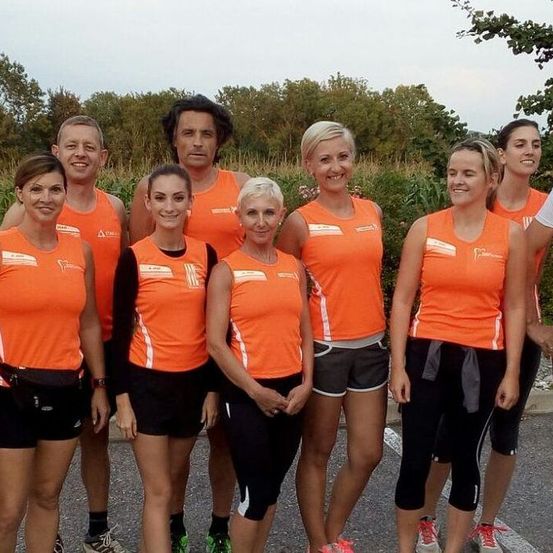 Eine Gruppe von Menschen in orangefarbener und schwarzer Sportbekleidung steht lächelnd für ein Foto auf einer Straße. Der Hintergrund zeigt Bäume und Grün.