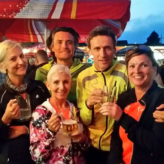 Fünf Personen in Jacken und mit Lächeln, halten Biergläser unter einem roten Regenschirm bei einer Veranstaltung im Freien.