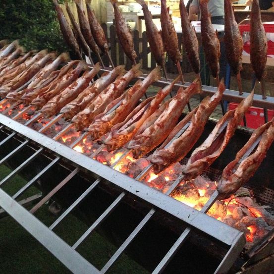 Ein großer Grill mit vielen Fischspießen darauf. Die Fische werden über offenem Feuer gegart, und der Grill ist aus Metall.