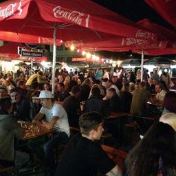 Ein Outdoor-Bereich bei Nacht mit vielen Menschen, die unter roten Coca-Cola-Sonnenschirmen sitzen. Die Tische sind mit Gläsern und Getränken gefüllt, und der Bereich ist hell beleuchtet.