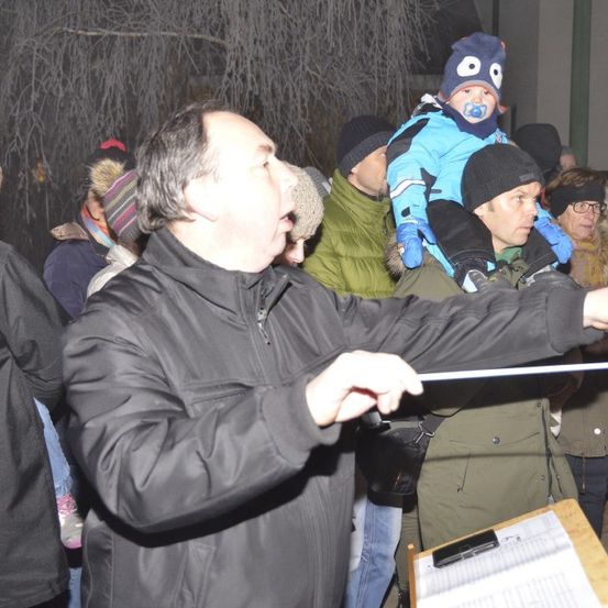 Ein Dirigent führt einen Chor bei Nacht im Freien an; ein Kind mit Schnuller sitzt auf seinen Schultern.