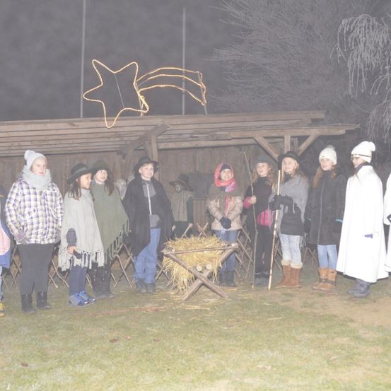 Eine Gruppe von Menschen steht draußen in einer Krippe, einige in Winterkleidung. Im Hintergrund steht eine Holzkrippe mit Heu darin.