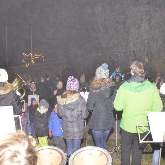 Eine Gruppe von Menschen in Winterkleidung versammelt sich nachts im Freien. Sie befinden sich in der Nähe eines Gebäudes mit einem sternförmigen Licht und Musikinstrumenten.