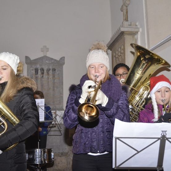 Drei Mädchen spielen Instrumente in einer Kirche. Sie tragen Winterkleidung und Mützen. Eine spielt Trompete, eine Tuba und das dritte ein weiteres Blechblasinstrument.