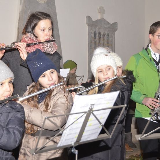 Eine Gruppe von Kindern und einem Erwachsenen spielt Musikinstrumente, darunter Flöten und eine Saxophon, in einem Raum mit einer Steinwand und einem Kreuz.