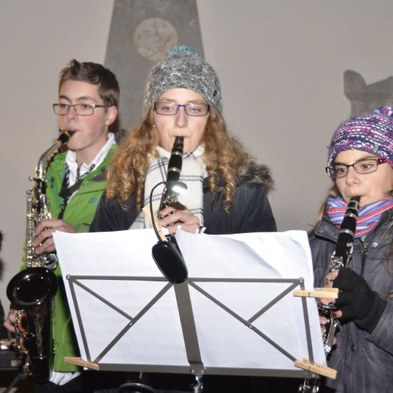 Drei Personen spielen Blasinstrumente in einem Raum. Die Person auf der linken Seite spielt eine Saxophon, die mittlere Person spielt eine Klarinette und die rechte Person ebenfalls eine Klarinette. Sie stehen hinter einem Notenständer.