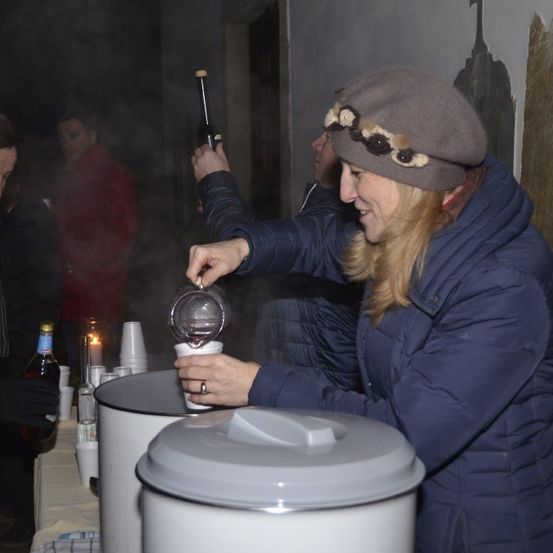 Eine Frau in einem blauen Mantel gießt ein Getränk in einen Becher. Hinter ihr stehen andere, einer hält eine Flasche. Die Szene ist schwach beleuchtet, mit Bechern und Flaschen auf einem Tisch im Hintergrund.