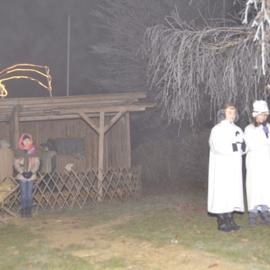 Zwei Personen in weißen Gewändern stehen vor einer kleinen Holzstruktur, wahrscheinlich einer Krippe, in einer nebligen Außenumgebung bei Nacht.