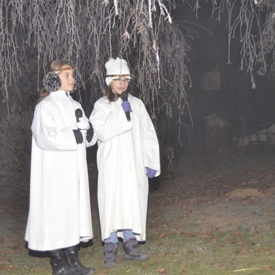 Zwei Frauen in weißen Gewändern stehen draußen und halten Mikrofone. Sie stehen unter einem Baum mit vereisten Ästen.