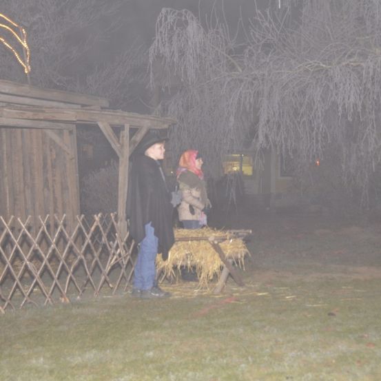 Zwei Menschen in Kostümen stehen in einem schwach beleuchteten Außenbereich, möglicherweise eine Krippe mit Heu auf einer Holzstruktur, umgeben von einem Holzzaun und Bäumen im Hintergrund.