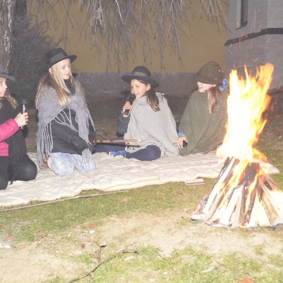 Vier Mädchen in Hüten sitzen um ein Lagerfeuer und halten Mikrofone. Ein Plaid liegt auf dem Rasen und ein Haus mit einem Fenster ist im Hintergrund.