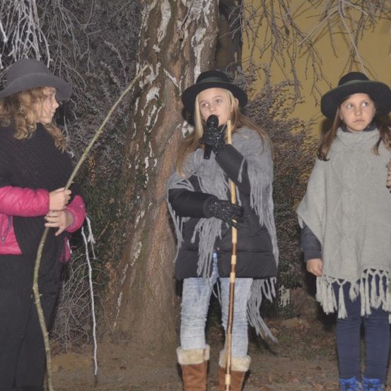 Drei Mädchen stehen bei einem Baum im Winter, gekleidet in Hüte, Schals und Stiefel. Ein Mädchen hält einen Stock und ein Mikrofon, ein anderes einen Stock und das dritte steht neben ihnen.