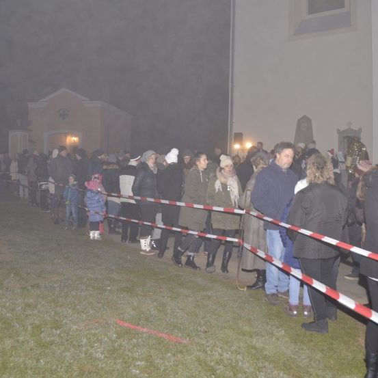 Menschen stehen in einer Reihe auf einem Rasenfeld vor einer Kirche und tragen Winterkleidung. Barrieren trennen die Schlange.