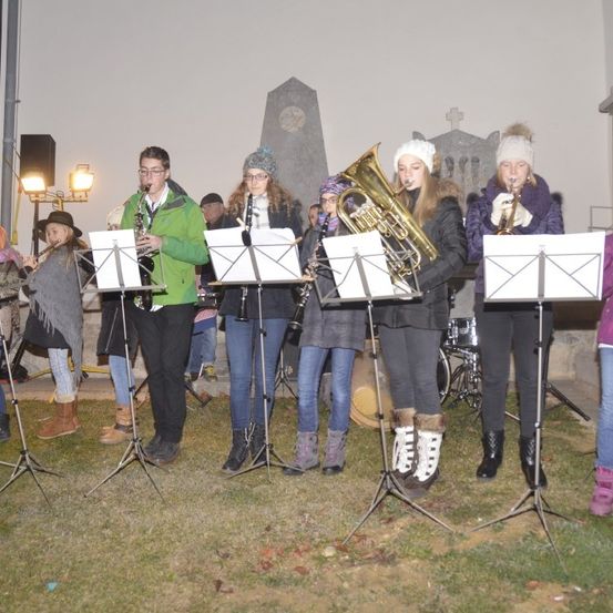Eine Gruppe von Kindern und Erwachsenen führt ein musikalisches Konzert im Freien auf und spielt verschiedene Instrumente wie Trompeten, Posaunen und Saxophone.