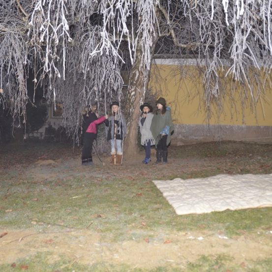 Vier Menschen in Winterkleidung stehen unter einem frostbedeckten Baum. Ein Teppich liegt auf dem Boden. Im Hintergrund befindet sich ein Gebäude.
