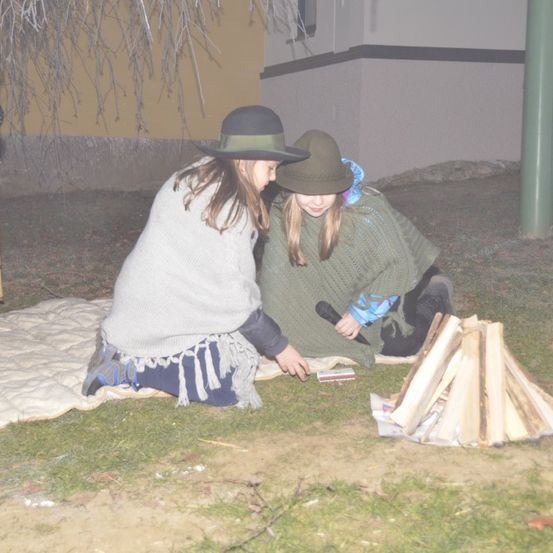 Zwei Mädchen sitzen auf einer Decke, eine hält ein Mikrofon und die andere schaut sich einen Feuerzeug in der Nähe eines Holzstapels an.