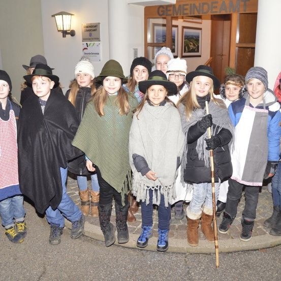 Eine Gruppe von Kindern in Winterkleidung steht vor einem Gebäude, wahrscheinlich einer Schule, und posiert für ein Foto. Sie tragen Mützen, Schals und Stiefel. Eine Lampe ist an der Wand hinter ihnen montiert.