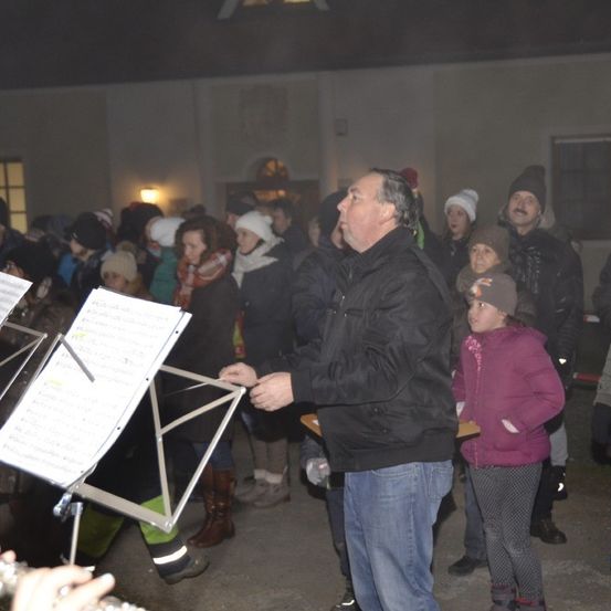 Ein Mann leitet einen Chor, mit einem Notenständer und Musiknoten vor ihm, während sich Menschen versammeln, einige in Winterkleidung.
