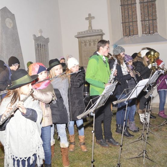 Eine Gruppe von Kindern und Erwachsenen steht vor einer Kirche und spielt Musikinstrumente wie Saxophon und Flöte auf einem Grasbereich.