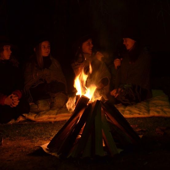 Vier Frauen sitzen um ein Lagerfeuer herum, das sie erhellt. Jede hält einen Becher, vielleicht für Wärme oder ein Getränk. Das Feuer brennt hell und wirft Schatten auf ihre Gesichter.